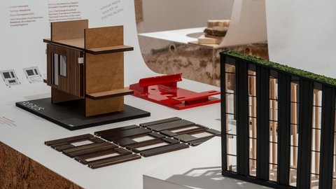 "Detailansicht eines Ausstellungstisches mit mehreren Architekturmodellen und Materialproben. Ein kleines Gebäude aus Holzplatten steht auf einem schwarzen Sockel mit der Aufschrift „REBUS“. Daneben liegen einzelne Fassadenelemente und ein Modell mit dunkler Außenhülle und begrüntem Dach. Im Hintergrund sind erklärende Texte und Grafiken auf weißen Tafeln zu sehen."
