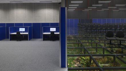 "Innenraum mit Reihen von schwarzen Stühlen auf einem transparenten Glasboden. Unter dem Glas sind Pflanzen, Erde und herabgefallene Blätter sichtbar. Seitlich befinden sich blaue Trennwände mit Arbeitsplätzen und Monitoren."