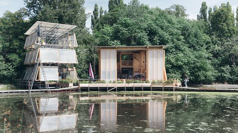 Zwei moderne Holzhäuser auf einem Steg über einem Teich mit Spiegelung und Bäumen im Hintergrund