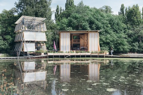 Zwei moderne Holzhäuser auf einem Steg über einem Teich mit Spiegelung und Bäumen im Hintergrund