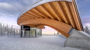 Geschwungene Holzkonstruktion einer Rennschlittenbahn mit verschneiter Umgebung und Glasgebäude darunter.
