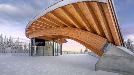 Geschwungene Holzkonstruktion einer Rennschlittenbahn mit verschneiter Umgebung und Glasgebäude darunter.
