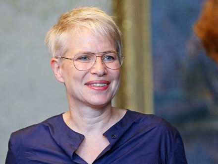 Lächelnde Person mit kurzem, hellem Haar und Brille in dunkelblauer Bluse vor unscharfem Hintergrund.