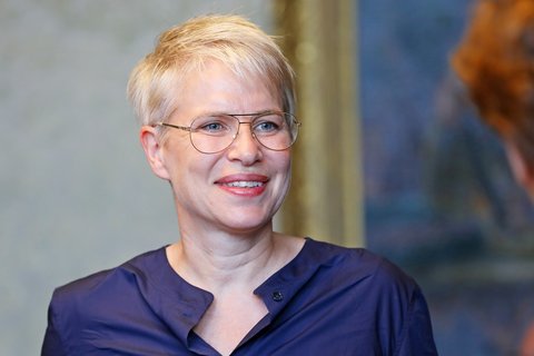 Lächelnde Person mit kurzem, hellem Haar und Brille in dunkelblauer Bluse vor unscharfem Hintergrund.