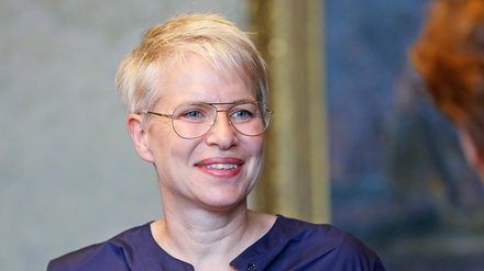 Lächelnde Person mit kurzem, hellem Haar und Brille in dunkelblauer Bluse vor unscharfem Hintergrund.