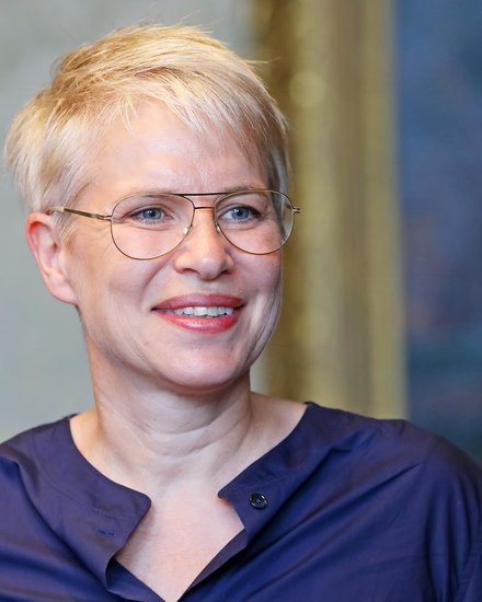 Lächelnde Person mit kurzem, hellem Haar und Brille in dunkelblauer Bluse vor unscharfem Hintergrund.