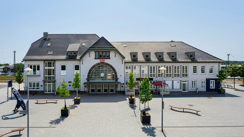 Historischer Bahnhofsvorplatz  Ein symmetrisch aufgebautes Bahnhofsgebäude mit hellem Putz, Satteldach und einem zentralen Eingang mit Uhr und DB-Logo. Davor liegt ein weiter, gepflasterter Vorplatz mit Bänken, jungen Bäumen in Pflanzkübeln und einzelnen Skulpturen. Der Platz wirkt ruhig und offen. Das Bild zeigt einen Bahnhof als städtebaulichen Mittelpunkt. Es steht für Ankunft, Abfahrt und den Bahnhof als sozialen und öffentlichen Raum.