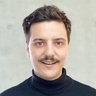 Porträt eines Mannes mit dunklem Haar und Schnurrbart vor hellem Hintergrund.