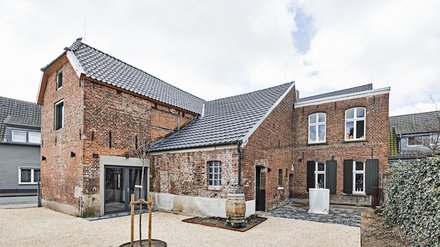 Innenhofansicht mehrerer ehemaliger Stalls- oder Scheunengebäude aus rotem Backstein mit modernen Türen, Kiesplatz und Fassdeko in heller, gepflegter Umgebung.