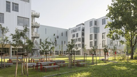 Moderner Gebäudekomplex mit mehreren Stockwerken und Außenbereich mit Bänken, Tischen und jungen Bäumen