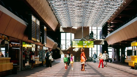Farbfoto einer weitläufigen Terminalhalle aus den 1970ern. Ein filigranes Raumfachwerkdach lässt Licht durch und wirft geometrische Schatten auf den Fliesenboden. Links sind Verkaufsstände mit gelben Theken und dunklen Anzeigetafeln. Mehrere Menschen in zeittypischer Kleidung bewegen sich durch den Raum, darunter eine Gruppe Kinder in der Mitte und eine Frau in einem roten Mantel. Grüne Hinweisschilder hängen von der Decke.