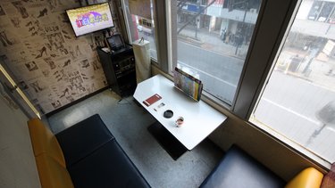 Kleine Karaoke-Box mit Sofa, Tisch und Bildschirm am Fenster über einer belebten Straße.