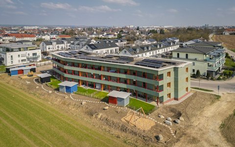 „Luftbild eines modernen, geschwungenen Wohngebäudes mit grüner Fassade, roten Balkonen und Solaranlage am Rande eines Neubaugebiets.“