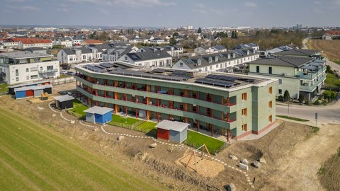 „Luftbild eines modernen, geschwungenen Wohngebäudes mit grüner Fassade, roten Balkonen und Solaranlage am Rande eines Neubaugebiets.“