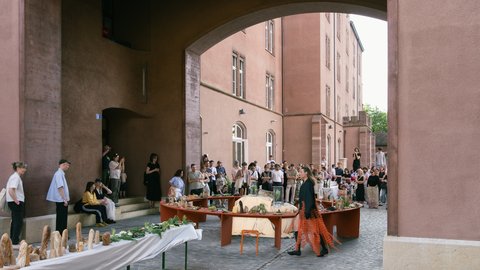 Eine große Menschenmenge versammelt sich in einem gepflasterten Innenhof unter einem massiven Torbogen. Im Zentrum steht eine kreisförmige Installation aus Holztischen, auf denen Objekte ausgestellt sind. Im Vordergrund ist ein langer, weiß gedeckter Tisch mit verschiedenen Brotskulpturen zu sehen.