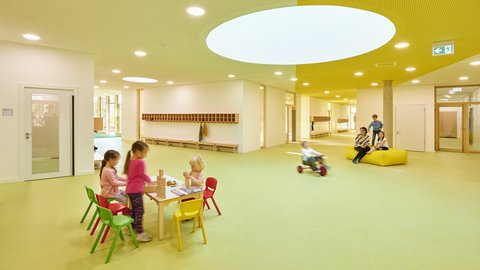 Kinder sitzen an einem kleinen Tisch mit bunten Stühlen in einem hellen, modernen Innenraum mit rundem Oberlicht