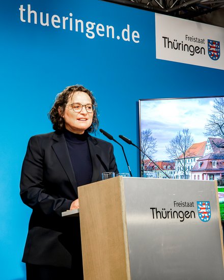 Person steht an einem Rednerpult mit dem Schriftzug 'Freistaat Thüringen' vor blauem Hintergrund und einem Bildschirm, der ein historisches Gebäude zeigt.