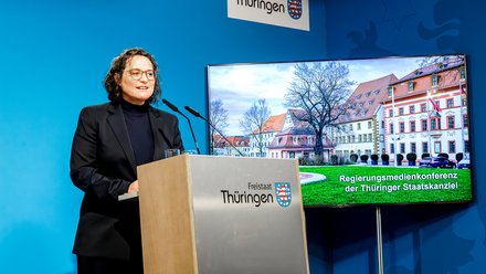 Person steht an einem Rednerpult mit dem Schriftzug 'Freistaat Thüringen' vor blauem Hintergrund und einem Bildschirm, der ein historisches Gebäude zeigt.