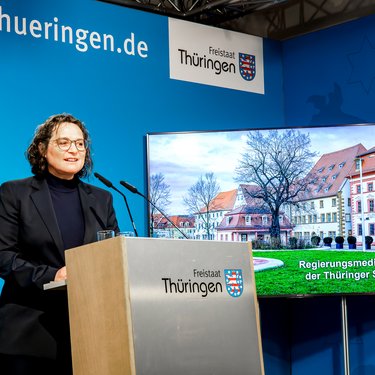 Person steht an einem Rednerpult mit dem Schriftzug 'Freistaat Thüringen' vor blauem Hintergrund und einem Bildschirm, der ein historisches Gebäude zeigt.