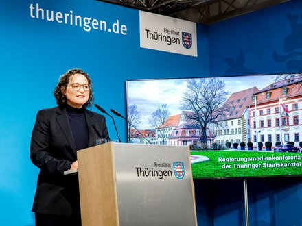 Person steht an einem Rednerpult mit dem Schriftzug 'Freistaat Thüringen' vor blauem Hintergrund und einem Bildschirm, der ein historisches Gebäude zeigt.