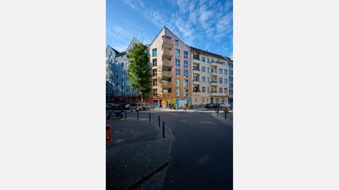 Straßenecke mit neuem Wohnhaus in Holzoptik und Balkonen zwischen hellen Putzbauten; breite Kreuzung mit Pollern, Rädern und parkenden Autos.