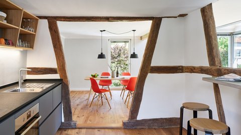 "Offene Wohnfläche in einem Fachwerkhaus mit massiven dunklen Holzbalken. Links schwarze moderne Küchenzeile mit Edelstahlgeräten. Rechts Essbereich mit Holztisch und roten Stühlen. Heller Holzboden, weiße Wände, große Fenster, schwarze Pendelleuchten. Der Raum verbindet historische Bausubstanz mit modernem Design."