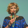 Frau mit kurzem lockigem Haar hält Mikrofon und gestikuliert mit ausgestrecktem Arm vor einer Leinwand mit unscharfem Text