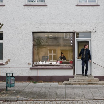Schaufensterbüro an Straßenecke: Fenster mit „architektur+textil“, Tür mit „co:büro“. Eine Person tritt heraus, innen sitzt jemand am Tisch. Kahl beschnittene Bäume, Straßenschild „Littelstraße“.