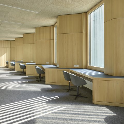 Langer Raum mit großen Fenstern, Holzverkleidungen und mehreren Arbeitsplätzen mit grauen Stühlen