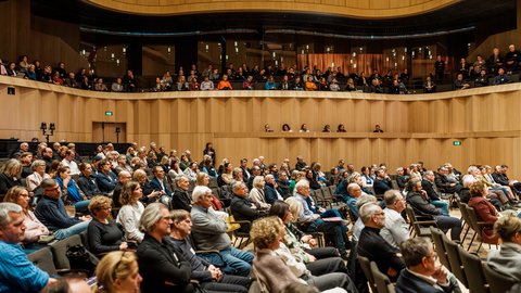 Großes Publikum sitzt in einem modernen Konzert- oder Veranstaltungssaal und hört einem Vortrag zu.