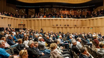Großes Publikum sitzt in einem modernen Konzert- oder Veranstaltungssaal und hört einem Vortrag zu.