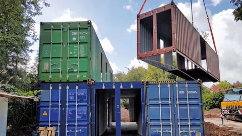 Mehrere übereinander gestapelte blaue und grüne Container, ein roter Container wird mit einem Kran gehoben, im Vordergrund Baustellengeräte und Erde