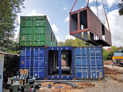 Mehrere übereinander gestapelte blaue und grüne Container, ein roter Container wird mit einem Kran gehoben, im Vordergrund Baustellengeräte und Erde