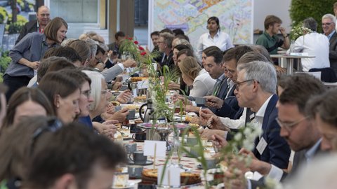 Lange Gemeinschaftstafel; viele Menschen essen, trinken und unterhalten sich.