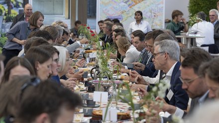 Lange Gemeinschaftstafel; viele Menschen essen, trinken und unterhalten sich.