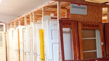 Reihe gebrauchter Zimmertüren in einem Lager: Türen stehen hochkant in Holzgestellen, überwiegend weiß, eine gelb; vorn eine große Holztür mit Glaseinsätzen. Ein Schild darüber lautet „Zimmertüren“.