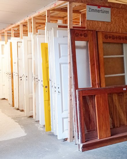 Reihe gebrauchter Zimmertüren in einem Lager: Türen stehen hochkant in Holzgestellen, überwiegend weiß, eine gelb; vorn eine große Holztür mit Glaseinsätzen. Ein Schild darüber lautet „Zimmertüren“.