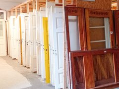 Reihe gebrauchter Zimmertüren in einem Lager: Türen stehen hochkant in Holzgestellen, überwiegend weiß, eine gelb; vorn eine große Holztür mit Glaseinsätzen. Ein Schild darüber lautet „Zimmertüren“.
