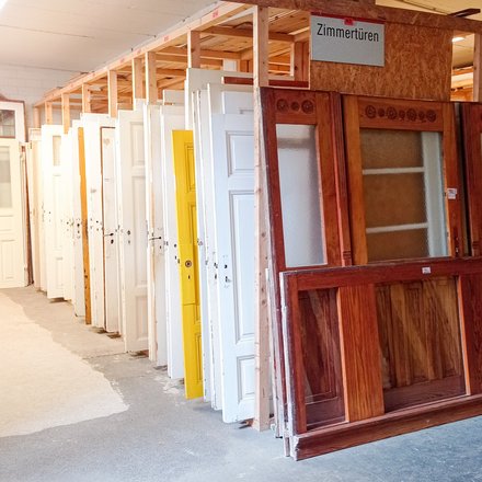 Reihe gebrauchter Zimmertüren in einem Lager: Türen stehen hochkant in Holzgestellen, überwiegend weiß, eine gelb; vorn eine große Holztür mit Glaseinsätzen. Ein Schild darüber lautet „Zimmertüren“.