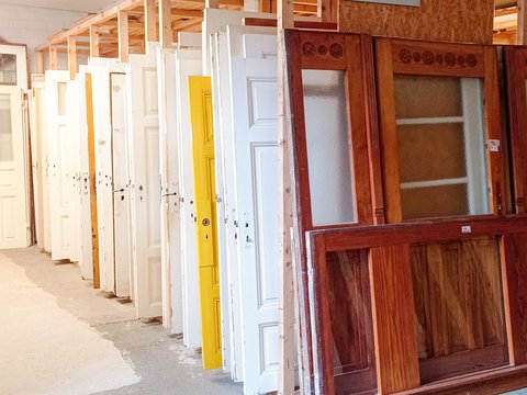 Reihe gebrauchter Zimmertüren in einem Lager: Türen stehen hochkant in Holzgestellen, überwiegend weiß, eine gelb; vorn eine große Holztür mit Glaseinsätzen. Ein Schild darüber lautet „Zimmertüren“.