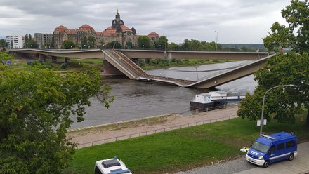Eine weitere Aufnahme zeigt die eingestürzte Brücke aus einer anderen Perspektive, einzelne Fahrbahnsegmente liegen geknickt über dem Flusslauf, Einsatzfahrzeuge stehen am Ufer, während das Umfeld abgesperrt ist, wodurch das Bild die akute Gefahrenlage und die Folgen des Bauwerksversagens dokumentiert und für die Notwendigkeit von Sicherheitsüberprüfung, langfristiger Planung und nachhaltiger Erneuerung von Infrastruktur steht.
