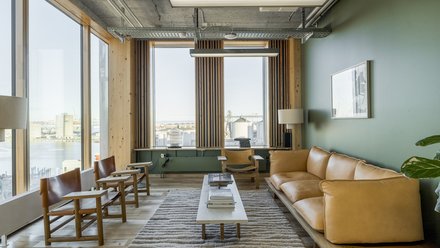 Heller, gemütlicher Lounge‑Raum mit großen Panoramafenstern zum Hafen: hellbraunes Leder‑Sofa rechts auf einem Holzpodest, links zwei Holzstühle mit Lederbespannung, in der Mitte niedriger Holztisch auf flauschigem Teppich. Holzvertäfelung und schlanke Lamellen rahmen die Fenster, die grüne Wand und eine Stehlampe sorgen für warme Akzente. Sichtbare Deckeninstallationen und eine große Hängeleuchte betonen die moderne, natürliche Materialität des Raums.