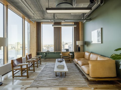 Heller, gemütlicher Lounge‑Raum mit großen Panoramafenstern zum Hafen: hellbraunes Leder‑Sofa rechts auf einem Holzpodest, links zwei Holzstühle mit Lederbespannung, in der Mitte niedriger Holztisch auf flauschigem Teppich. Holzvertäfelung und schlanke Lamellen rahmen die Fenster, die grüne Wand und eine Stehlampe sorgen für warme Akzente. Sichtbare Deckeninstallationen und eine große Hängeleuchte betonen die moderne, natürliche Materialität des Raums.