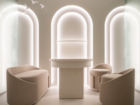  "Modernes Wartezimmer mit minimalistischem Design. Ein beige-weißes Farbschema mit drei gerundeten Sesseln in Beige-Tönen, die symmetrisch um einen weißen zylindrischen Tisch angeordnet sind. Im Hintergrund drei große bogenförmige Fenster mit integrierten LED-Leuchten. Deckenelemente mit weißen Vögeln. Die Beleuchtung schafft eine beruhigende, helle Atmosphäre."