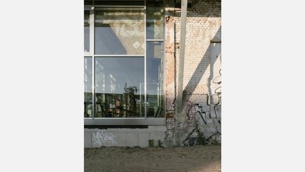 Außenwand: großes Glasfenster neben gealterter Ziegelwand mit Graffiti; Spiegelungen zeigen Hallenstruktur innen.