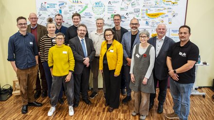 Eine Personengruppe steht im Halbkreis vor einem Whiteboard mit Blick in die Kamera 