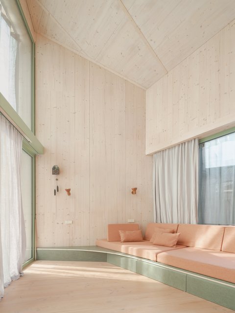 Heller Wohnraum mit Holzdecke und Wänden in Naturholztönen. Grüner Fensterrahmen mit weißen Vorhängen. Terracottafarbiges Sofa mit Kissen an grüner Sitzbank. Minimalistisches Design."