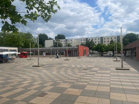 „Foto des aktuellen Zustands: Eine weitgehend versiegelte, gepflasterte Platzfläche mit Straßenlaternen, einem flachen roten Pavillon und Wohnblöcken im Hintergrund.“