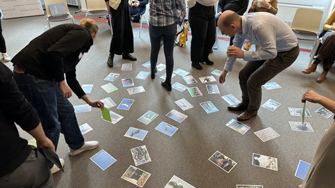 Blick von oben auf eine Gruppe von Menschen in einem Workshop. Auf einem grauen Teppichboden sind etwa 50 verschiedene kleine Fotos und Postkarten in einem lockeren Kreis verteilt. Die Teilnehmenden stehen um die Bilder herum, einige beugen sich hinunter oder halten Karten in der Hand, um sie auszuwählen oder zu betrachten. Im Hintergrund stehen Stühle im Kreis.
