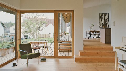 Moderner Wohnraum mit großen Fenstern, Holztreppe, grünem Sessel und Essbereich im Hintergrund